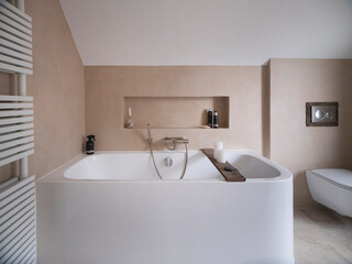 Fototapeta premium Contemporary bathroom featuring white bathtub with wooden bath caddy, beige walls, and modern heated towel radiator