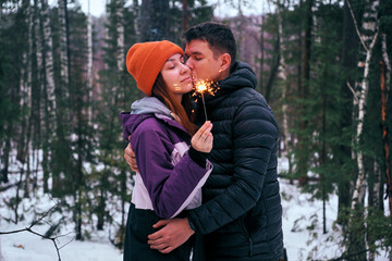 A young couple in a snowy forest is celebrating Christmas in the winter, holding a sparking sparkler. The concept of a cozy New Year and Christmas celebration with family