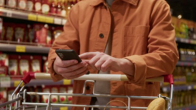 Midsection view of anonymous man wearing orange jacket using smartphone and leaning on handle of shopping cart inside grocery store