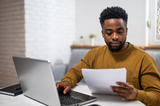 Concentrated African freelancer using laptop and examining documents while working from his cozy home.	