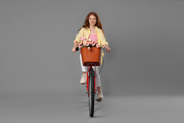 Beautiful teenage girl riding bicycle with basket of flowers against grey background