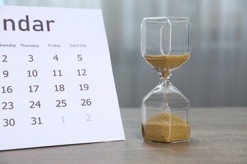 Deadline. Hourglass with flowing sand and calendar on wooden table indoors, closeup