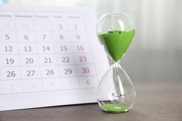 Deadline. Hourglass with flowing sand and calendar on wooden table indoors, closeup