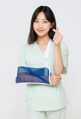 Young Asian woman in medical patient clothing with arm in a sling, smiling and making finger heart gesture. Concept of recovery, healthcare, support, and positive attitude during treatment. Studio sho