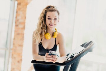 Active young woman with yellow headphones using smartphone on treadmill in gym, showcasing fitness,...