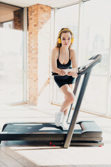 Young woman wearing headphones exercises on a treadmill in a bright gym, emphasizing fitness,...