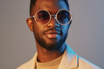 Young Black man wearing stylish round sunglasses, light grey suit jacket, and beige turtleneck,...