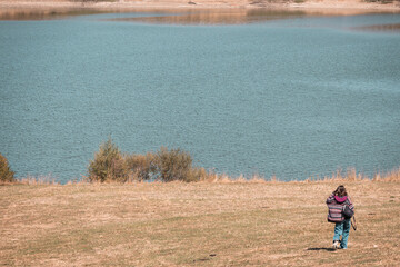 Young woman walking alone on dry grass toward a lake, surrounded by peaceful nature. Back view....