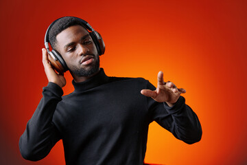 Young Black man enjoying music with headphones against a vibrant orange background, expressing joy...