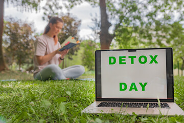 Close-up laptop screen DETOX DAY green text grass foreground woman seated reading book blurred...