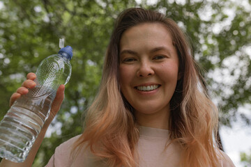 Smiling woman in pale shirt holding clear sport water bottle with blue cap standing outdoors under...