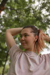 Naklejka premium Close-up view of woman adjusting hair into ponytail while smiling slightly, surrounded by natural light and green foliage. Casual moment showing readiness and calm presence.