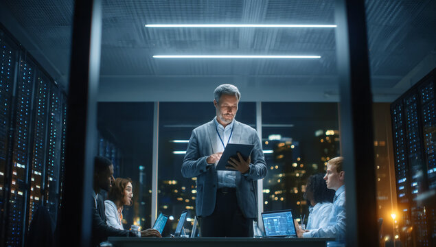 Tech team collaborates in data center at night. Leader reviews tablet. Modern, corporate look for web, marketing.