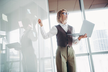 Fototapeta premium Confident businesswoman using a laptop and pointing at notes on a transparent board in a modern office