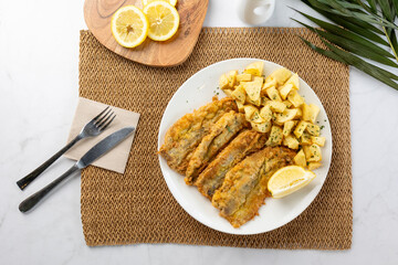 Breaded fried sardines with roasted potatoes and lemon slice on white plate