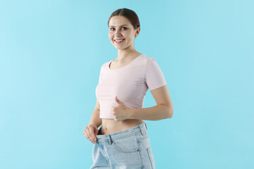 Beautiful woman wearing old jeans after weight loss and showing thumbs up on light blue background