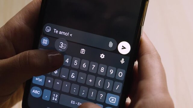 Close-up of a hand typing a love message in Spanish on a smartphone, typing "Te amo! <3" on a dark screen.