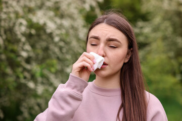 Woman with paper tissue suffering from seasonal pollen allergy near blossoming shrub on spring day