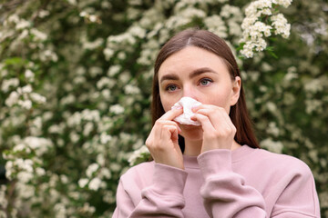 Woman with paper tissue suffering from seasonal pollen allergy near blossoming shrub on spring day,...
