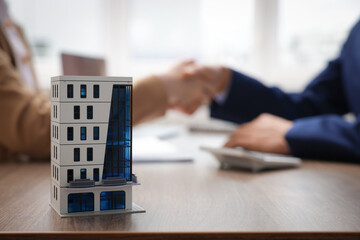 Real estate agent and client shaking hands at desk in office, focus on house model