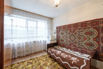 Cozy room with a couch against a carpet-covered wall. A window with sheer curtains lets in light. A...