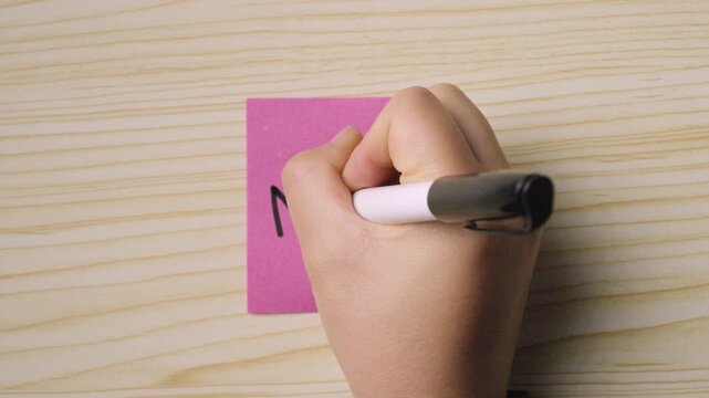 Handwriting the French word &ldquo;Merci!&rdquo; on a pink sticky note placed on a light wooden surface.