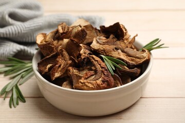 Pieces of dried mushrooms and rosemary in bowl on light wooden table, closeup