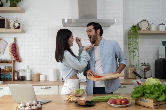 Young couple enjoying fun cooking moment in kitchen, woman feeding man with fork while he smiles playfully
