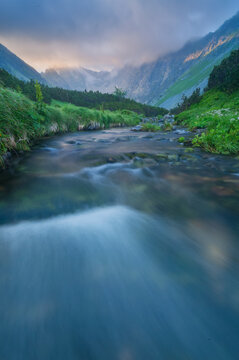 Fototapeta View of a serene river winding through lush greenery towards towering mountains under a cloudy sky, a tranquil escape, VysokÃ© Tatry, PreÅ¡ov Region, Slovakia.
