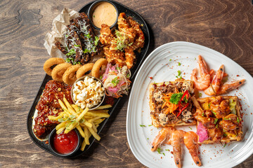 Assorted comfort foods including fried chicken, ribs, shrimp, onion rings, fries, and waffles, served on a wooden table in a savory and indulgent presentation.