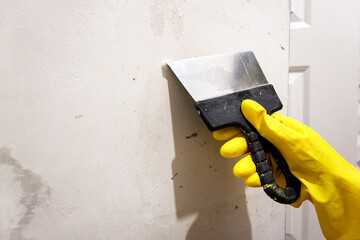 A hand in a yellow glove applies putty with a spatula to the wall