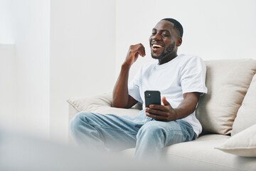 Young African man enjoying time at home, laughing while using smartphone, casual outfit,...