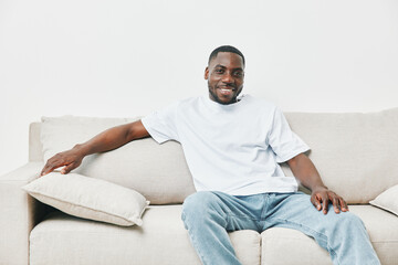 Cheerful young African American man in casual clothing relaxing on a beige sofa, conveying a sense...