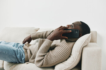 Relaxed young man in a cozy sweater holding a smartphone on a couch, exuding casual comfort and...