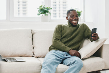 Smiling young Black man relaxing on a sofa, using a smartphone in a cozy, modern living room,...