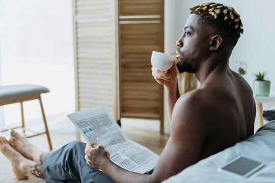 Side view of african american man with vitiligo holding coffee and newspaper in bedroom