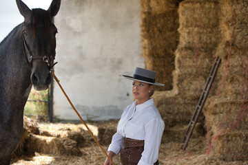 A young, beautiful woman dressed in riding clothes and a traditional Spanish hat is standing next...