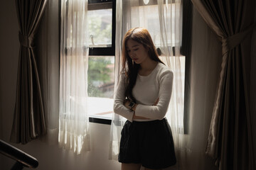 Sad and depressed young asian woman standing at window in the bedroom, Woman worry, Unhappy girl.