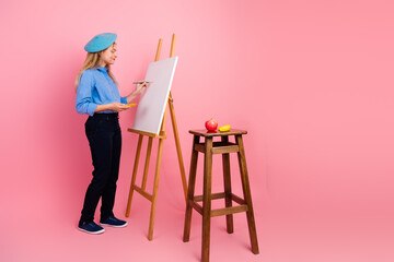 Fototapeta premium Young girl in artistic attire painting a still life canvas with fruits and stool on pink background