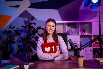 Young woman blogger with a book featuring a heart icon, seated in a modern office, surrounded by a...