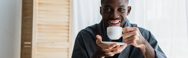 Positive african american man with vitiligo holding coffee cup and looking at camera at home, banner