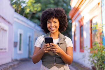 Smiling Afro-Brazilian woman using smartphone in colorful colonial street, representing travel...