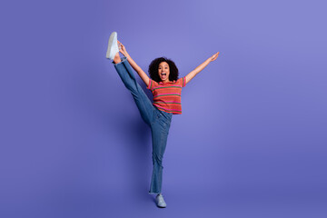 Fototapeta premium Energetic young woman in casual attire enthusiastically posing with raised leg on a vibrant purple background