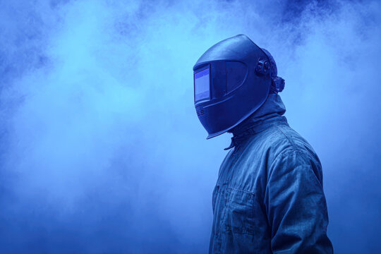 A person wearing a protective welding helmet and suit stands against a smoky blue background, suggesting industrial or welding work.