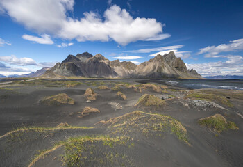 panorama of Vestrahorn mountains and black sand dunes topped with grass patches on a sunny summer...