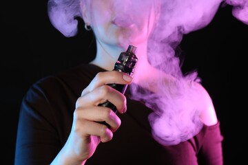Woman using electronic smoking device in neon lights on black background, closeup