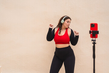 Curvy woman wearing sportswear listening music with headphones and recording fitness workout video with smartphone on tripod