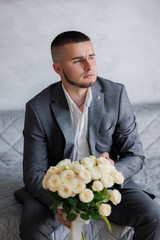 Young man in suit holding bouquet of flowers while sitting on grey sofa