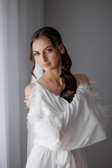 Elegant woman in white dress with feathers poses by a window in natural light