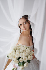 Bride posing gracefully with a bouquet of white roses behind sheer curtains